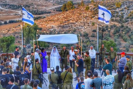 חופה בבסיס צבאי - רוב הקהל ואף החתן במדים, מצידי החופה הלבנה - דגלי ישראל מתנופפים ומעליה גרילנדות