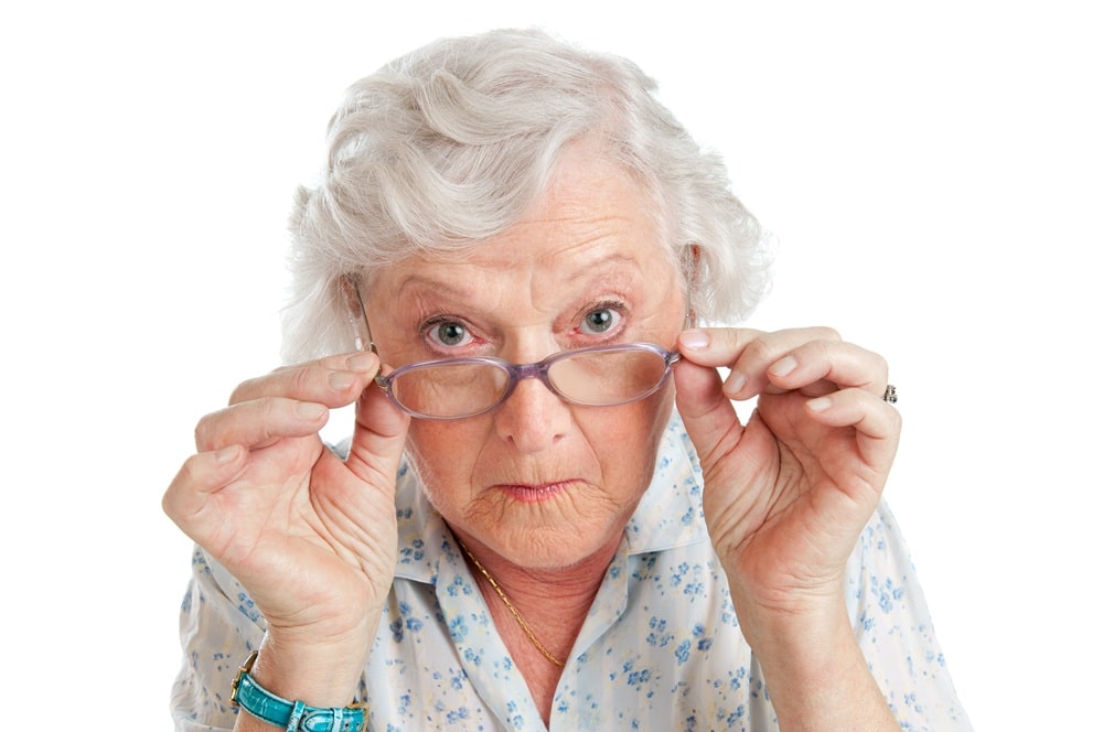אישה מבוגרת על רקע לבן מישירה מבט בוחן למצלמה וידיה אוחזות במשקפיים